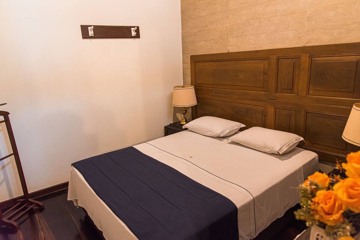 A hotel room with a wooden headboard, a bed with white bedding, a nightstand with a lamp, and a small table with a vase of flowers.