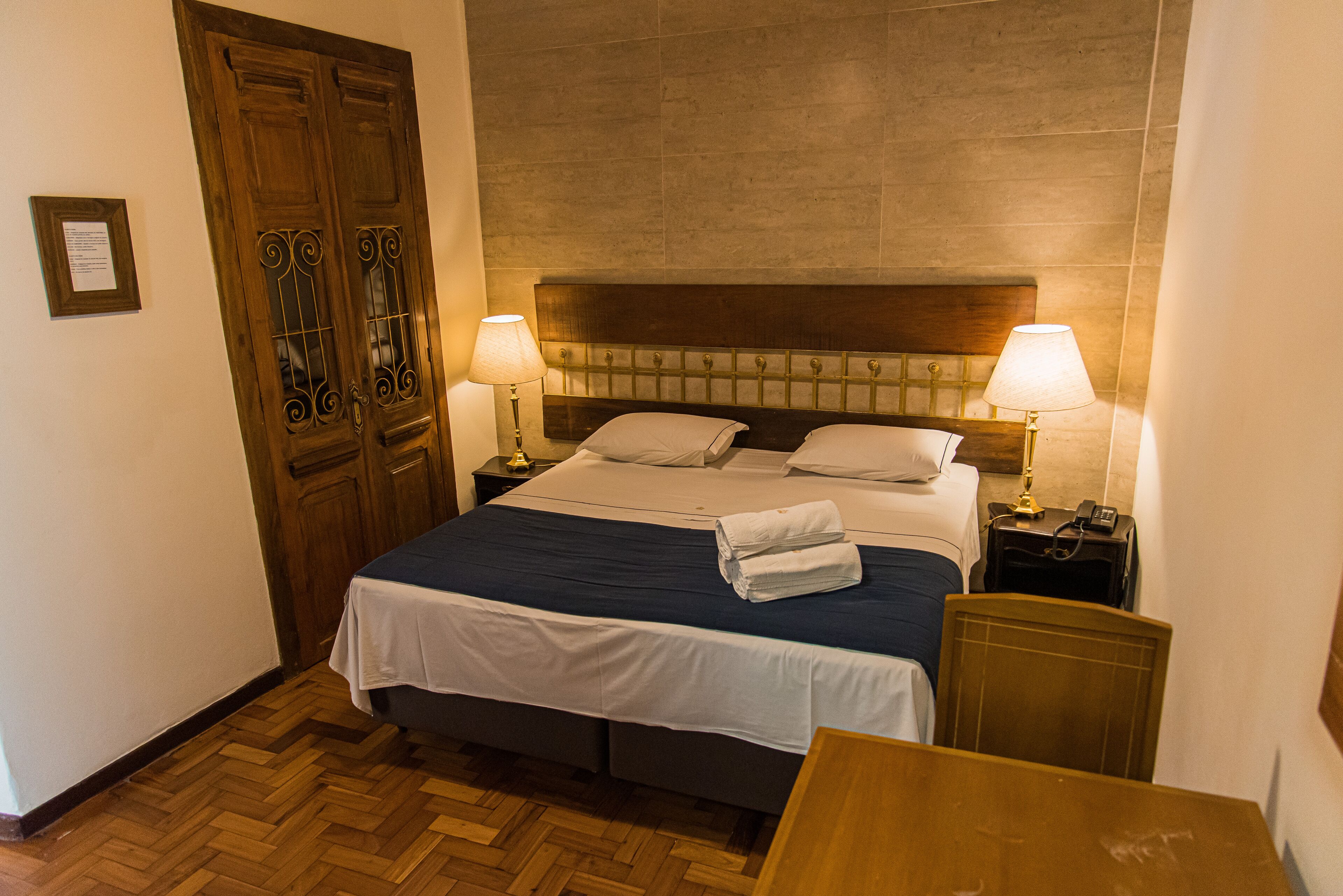 Quarto de hotel com uma cama grande, duas mesas de cabeceira, uma cabeceira de madeira e piso de madeira.