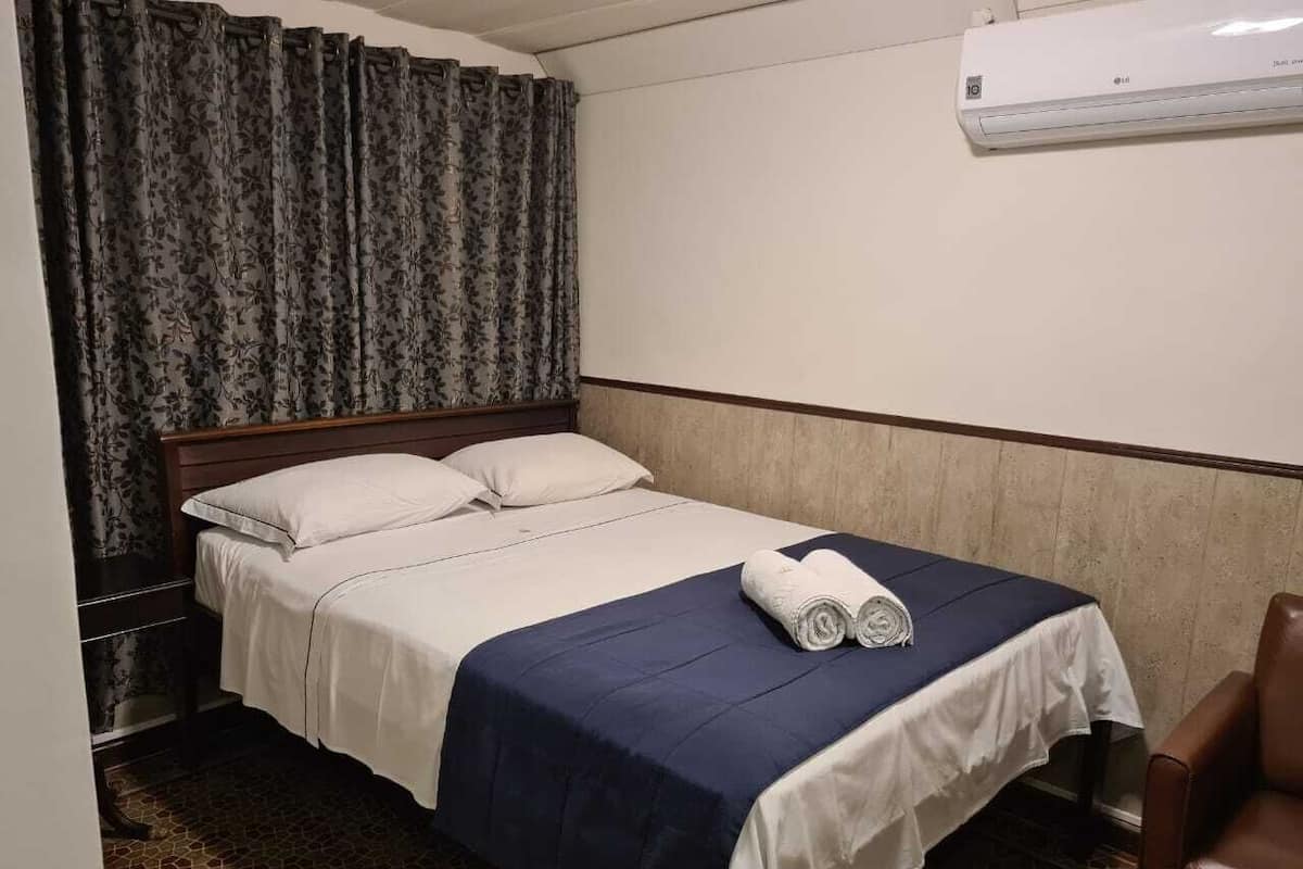 A hotel room with a bed, a table with towels, a chair, and an air conditioning unit.