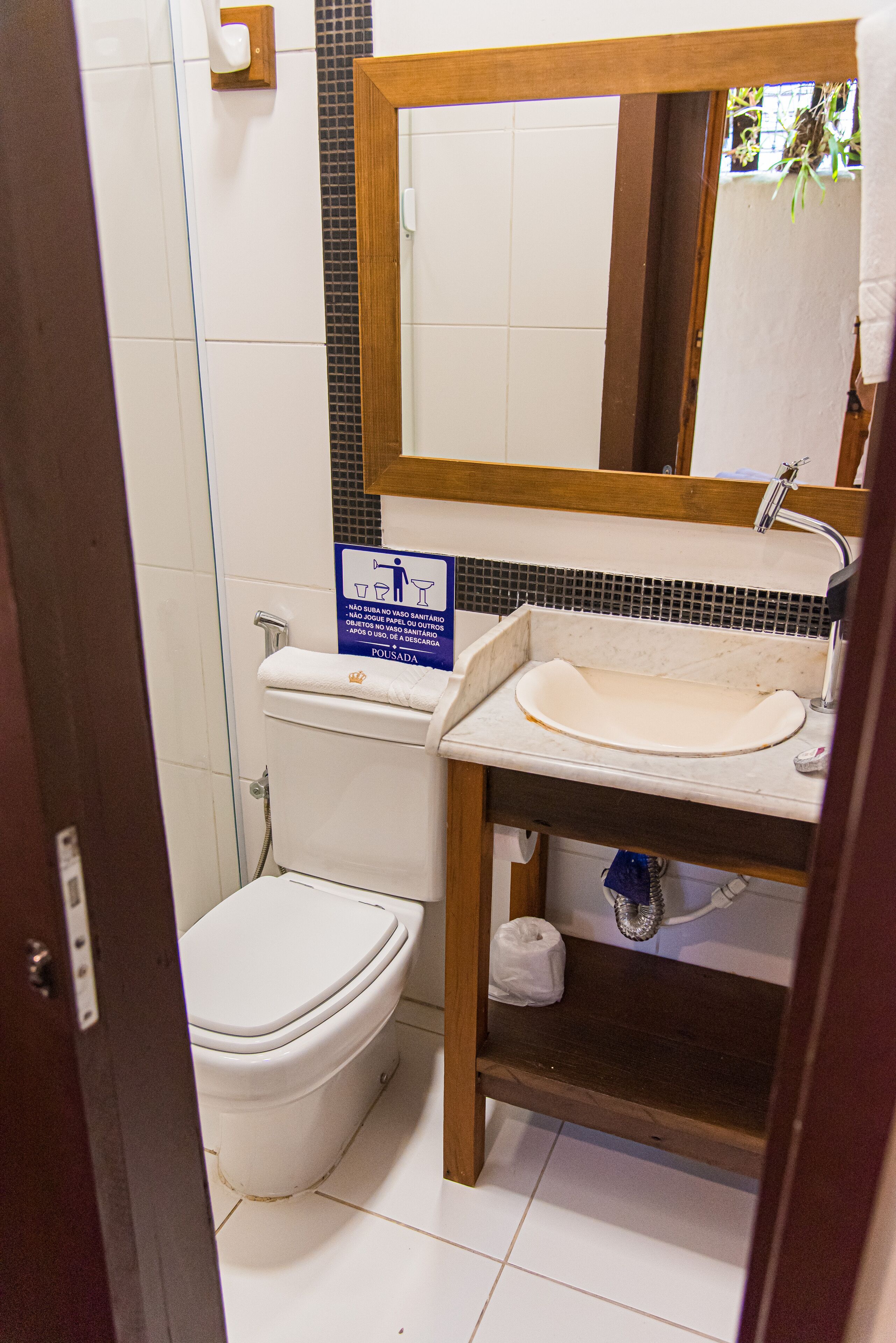 Une salle de bain avec des toilettes, un lavabo et un miroir. Il y a un panneau sur le mur et une serviette accrochée à la porte.