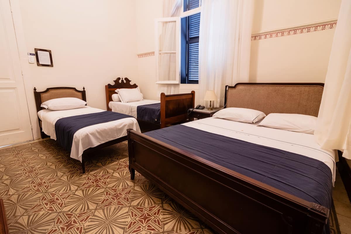 A hotel room with two single beds, a large bed, a patterned floor, and a window with curtains.