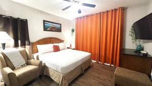 Une chambre d’hôtel avec un lit, deux fauteuils, un ventilateur de plafond et un tableau au mur.