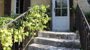 Una escalera de piedra conduce a una puerta blanca con un ventanal encima, rodeada de vegetación y una barandilla de hierro forjado.