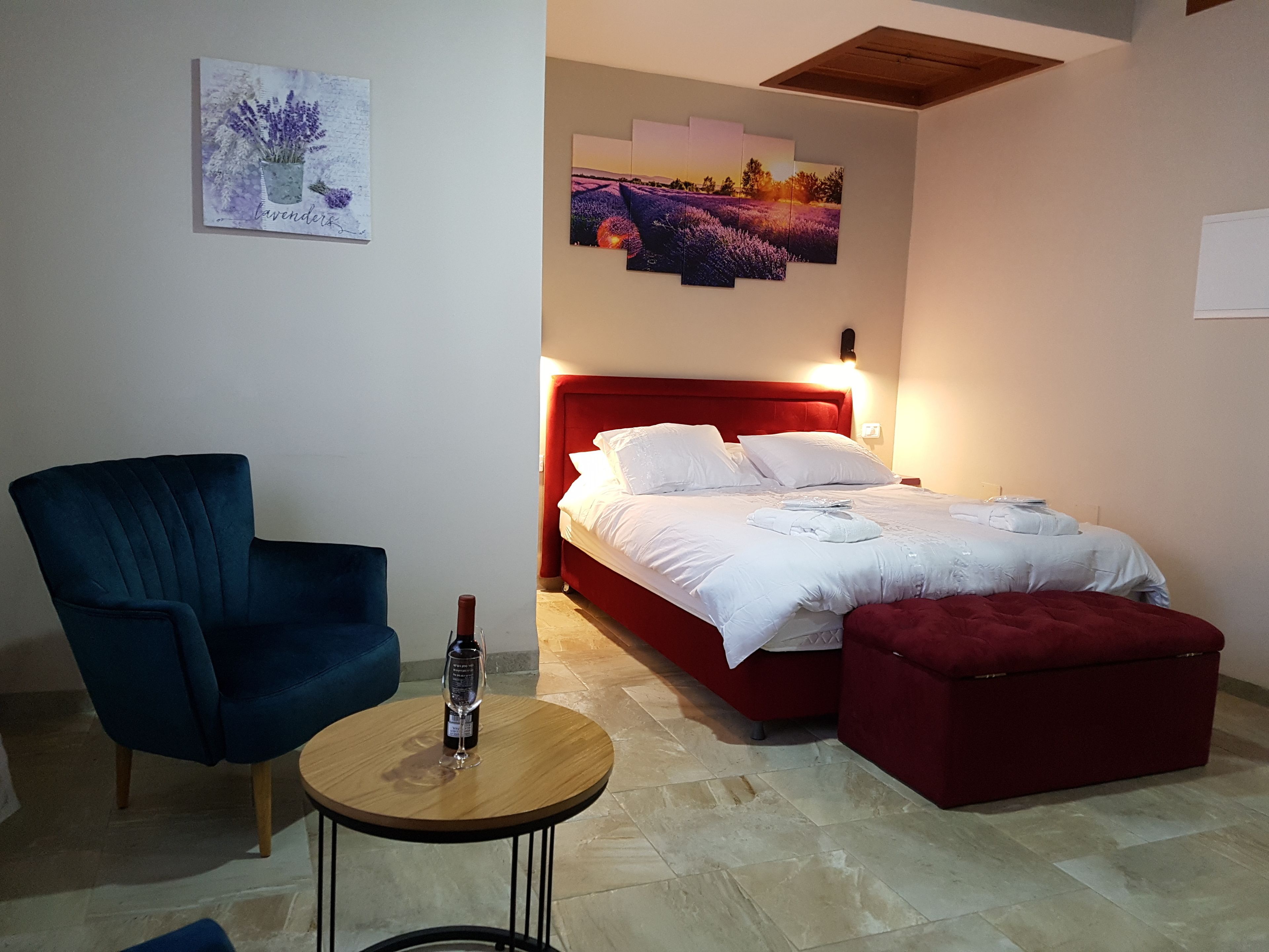 Een slaapkamer met een rood hoofdeinde, een bed met witte lakens, een blauwe fauteuil, een klein houten tafeltje met een wijnglas en een rode poef.