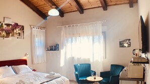 Un dormitorio con techo de madera, una cama con ropa blanca, un reposapiés rojo, dos sillas azules, una pequeña mesa redonda con una botella y vasos, y un televisor sobre un mueble.