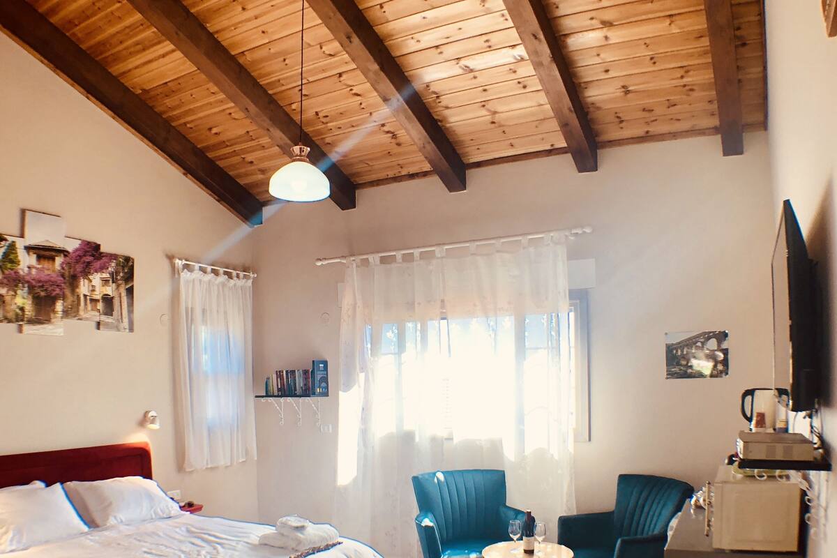 A bedroom with a wooden ceiling, a bed with white linens, a red ottoman, two blue chairs, a small round table with a bottle and glasses, and a television on a stand.