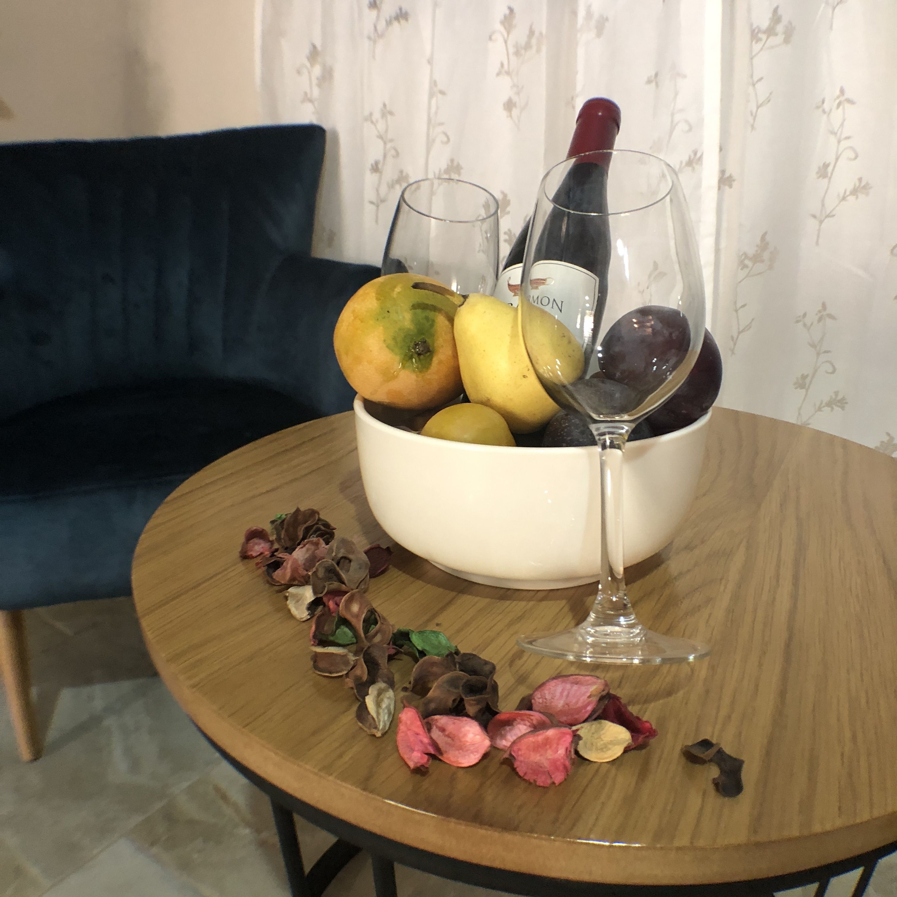 A round wooden table with a bowl of fruits and a wine bottle.