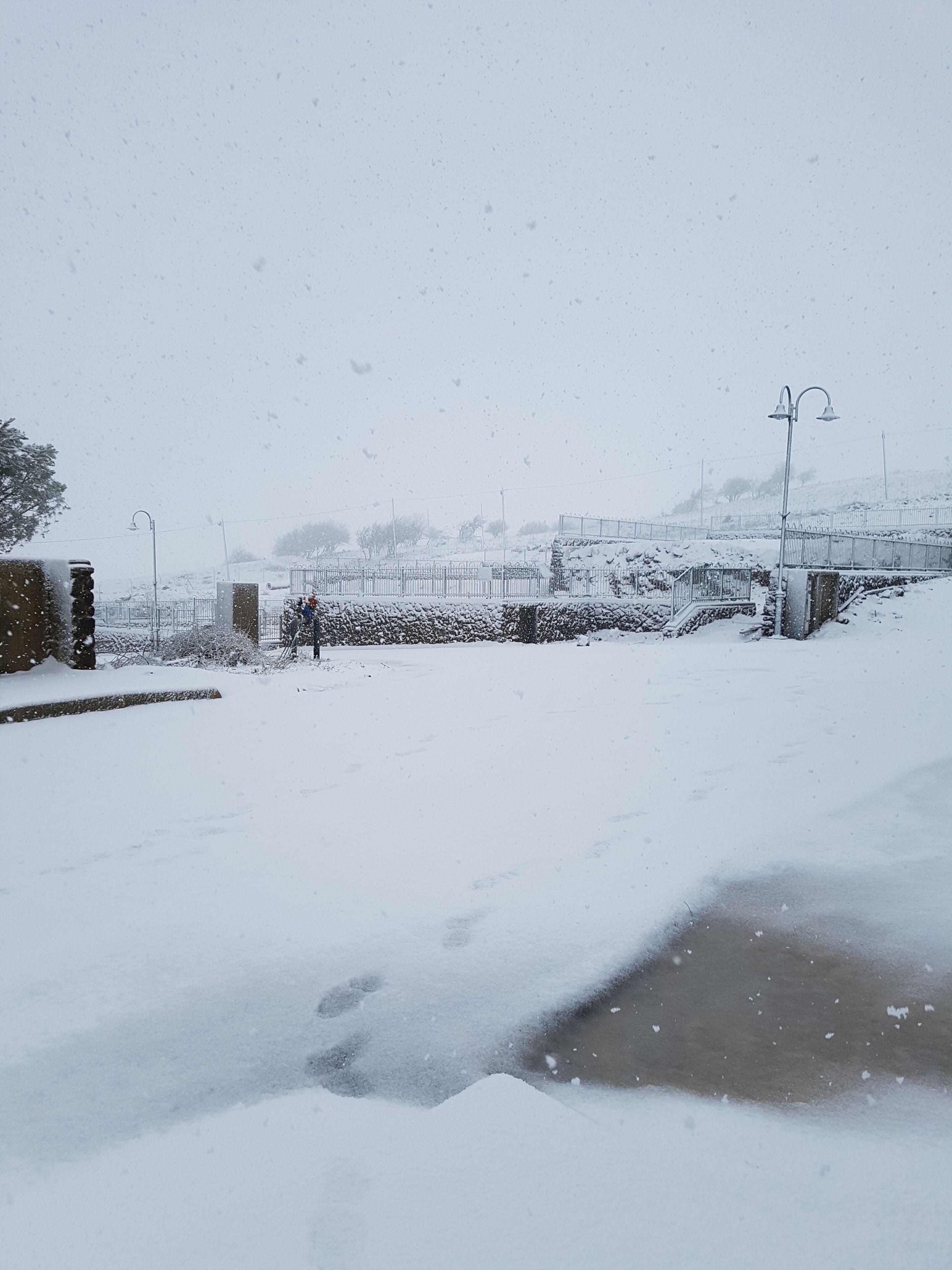 A snow-covered landscape with a stone wall, metal fence, and a bridge in the background.
