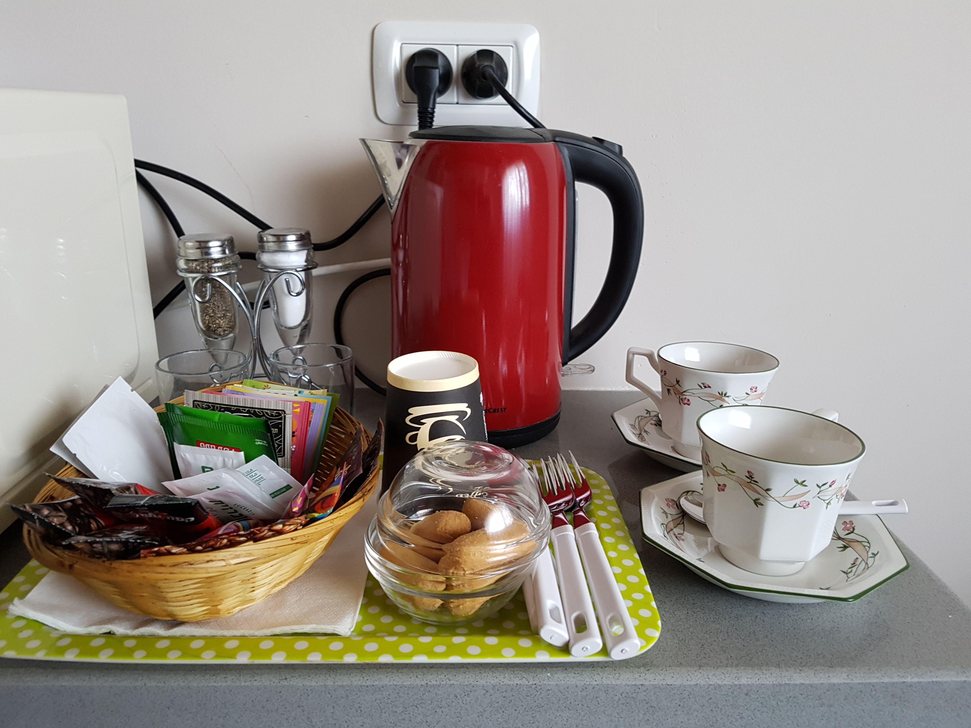 Una preparación para el desayuno con una tetera roja, tazas, una cesta de bocadillos y un tarro de cristal sobre un mantel de lunares verdes.