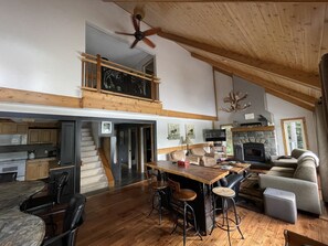 A spacious living area with a high ceiling, wooden beams, a dining table with bar stools, a fireplace, and a television.
