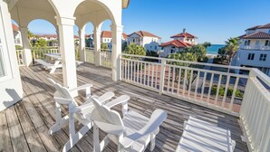 A wooden deck with white chairs and a table, overlooking a beach with buildings in the background. - Beach & Bay views! 4BR/3.5BA, Elevator, Com Pool/Tennis Court (St. George Island)