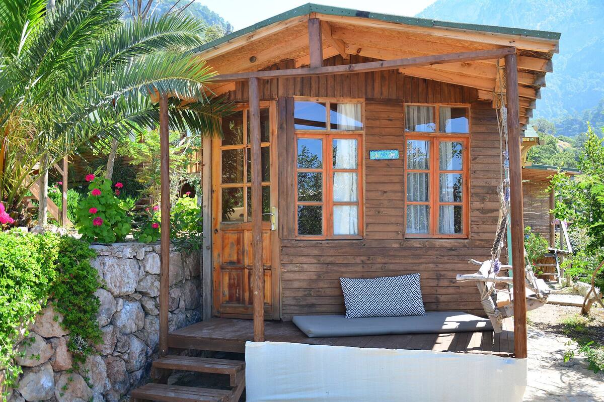 A wooden cabin with a porch, stone wall, and a bench with a pillow.
