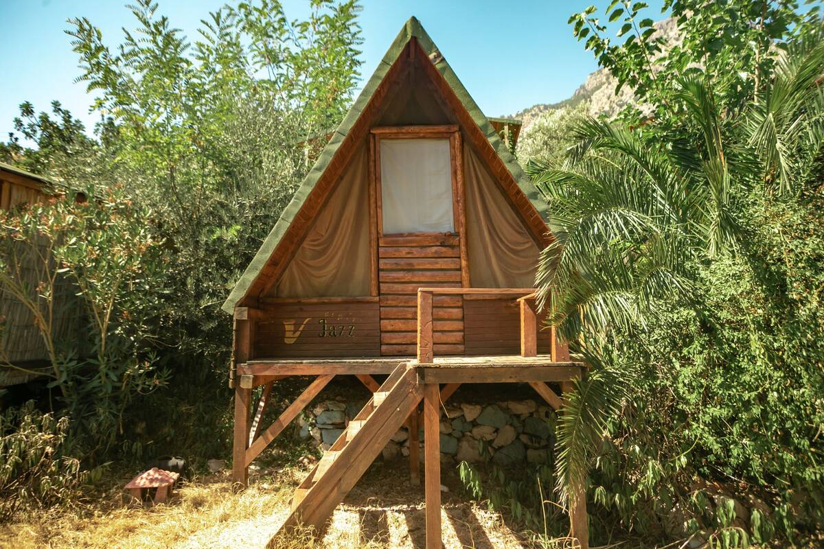 A wooden cabin with a balcony, surrounded by greenery and a mountain backdrop.