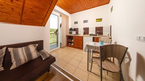 A cozy room with a wooden ceiling, a sofa with cushions, a dining table with chairs, a kitchen area with a microwave, and a sliding glass door leading to a balcony. - Szabolcs Apartments (Demjén)