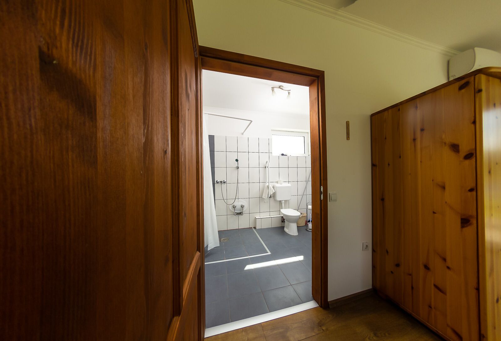 Une porte en bois donne accès à une salle de bain équipée de toilettes, d’une douche et d’un sol carrelé.