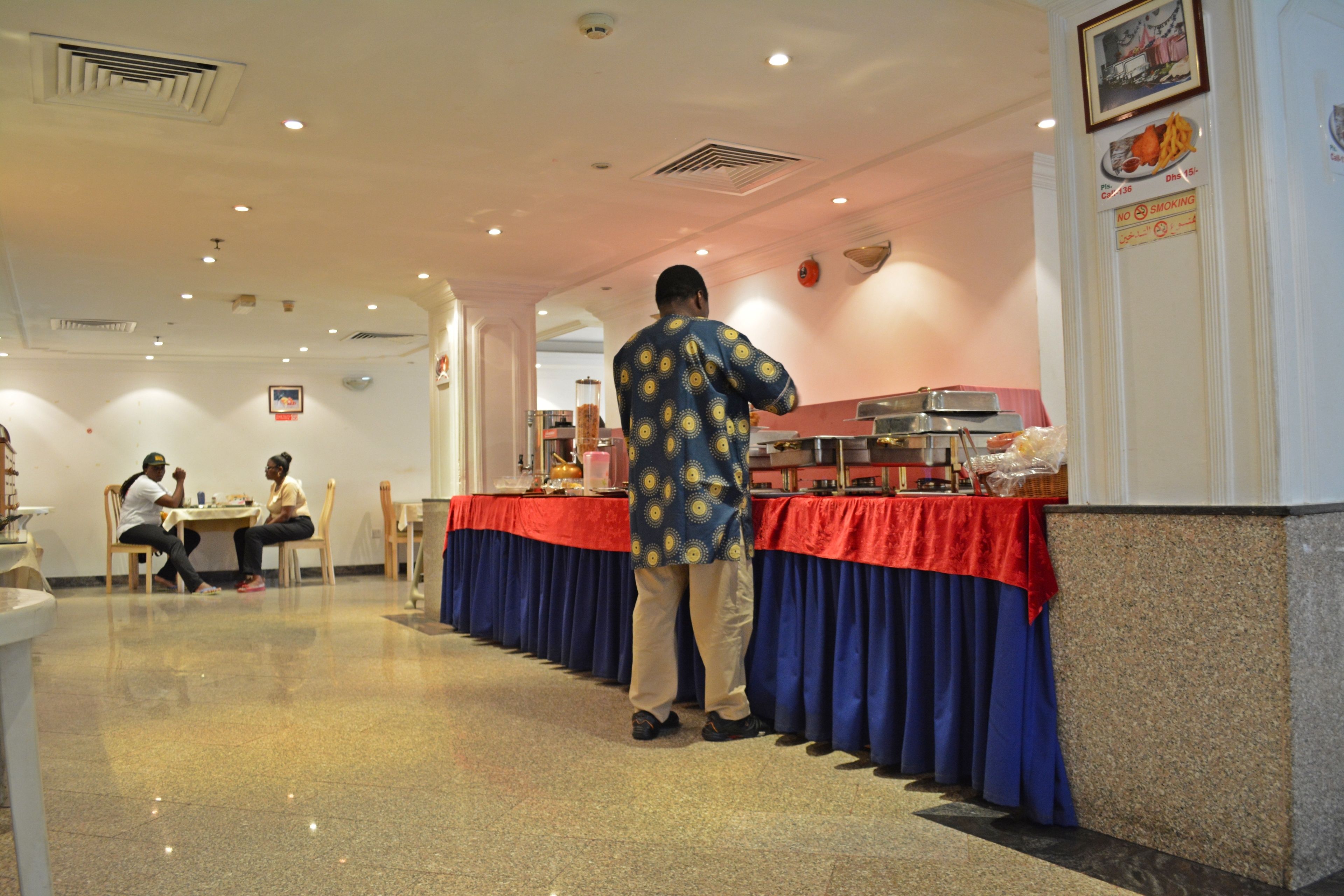 A buffet area with a long table covered in a red cloth, a man standing in front, and two people seated at a table in the background.