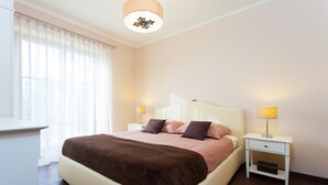 A modern bedroom with a large bed, two bedside tables with lamps, a desk, and a window with sheer curtains.
