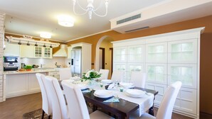 A dining area with a dark wooden table set for four, surrounded by white chairs. A chandelier hangs from the ceiling, and there is a large window with frosted glass panels on the right.