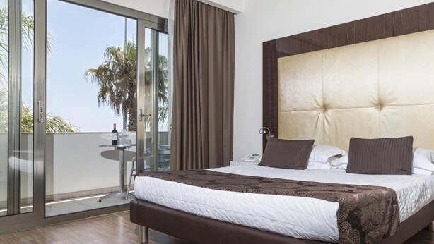 A modern bedroom with a large bed, a glass door leading to a balcony, and a view of palm trees. - La Bussola Hotel (Milazzo)