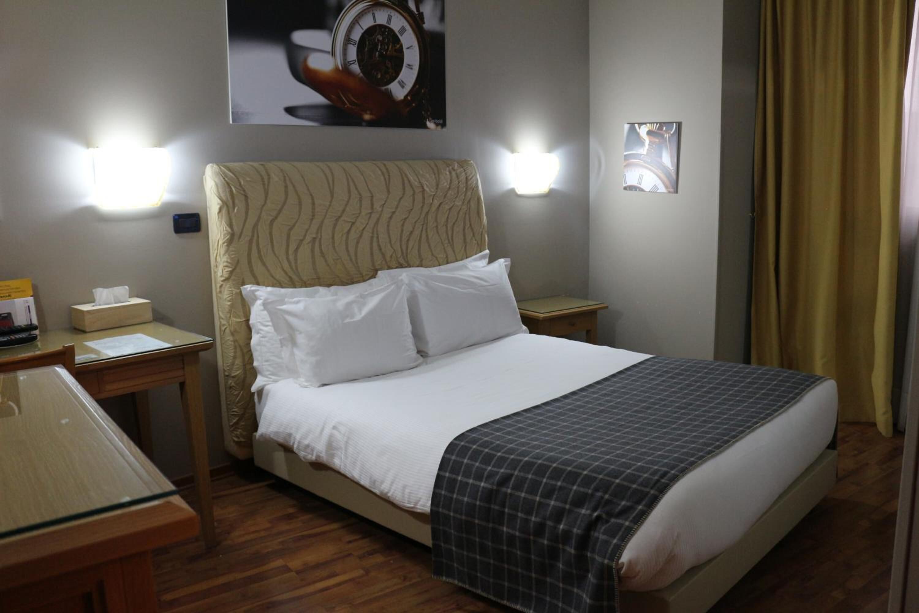 A hotel room with a bed, bedside tables, a desk, and a wall clock.