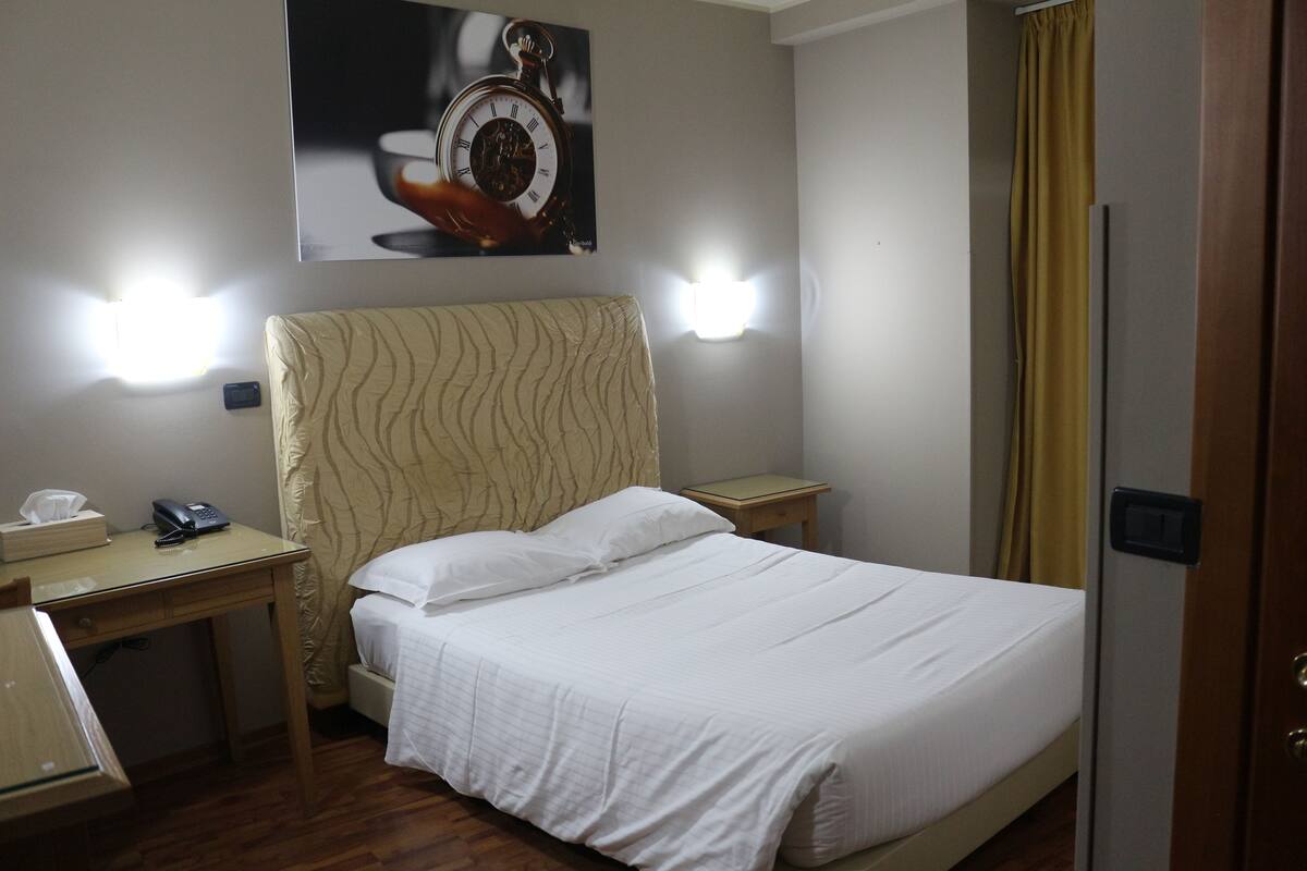 Quarto de hotel com cama, mesas de cabeceira, escrivaninha, telefone e relógio de parede.