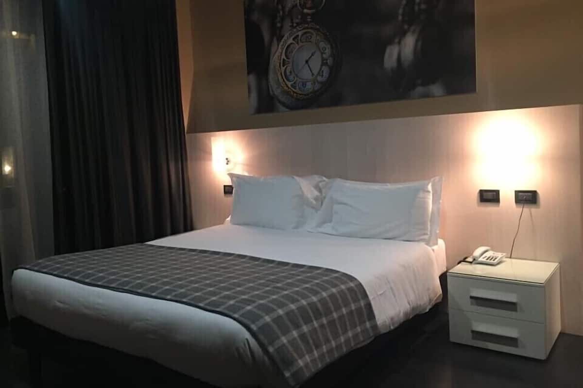 Quarto de hotel com uma cama grande, mesa de cabeceira com telefone e relógio fixado na parede.