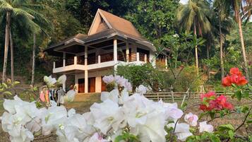 A two-story house with a balcony, surrounded by lush greenery and flowering plants.