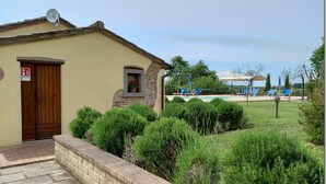 Ein Gebäude mit Holztür, Satellitenschüssel und einem kleinen Fenster mit Blumentopf. Im Hintergrund ist ein Poolbereich mit blauen Liegestühlen und Sonnenschirmen zu sehen.