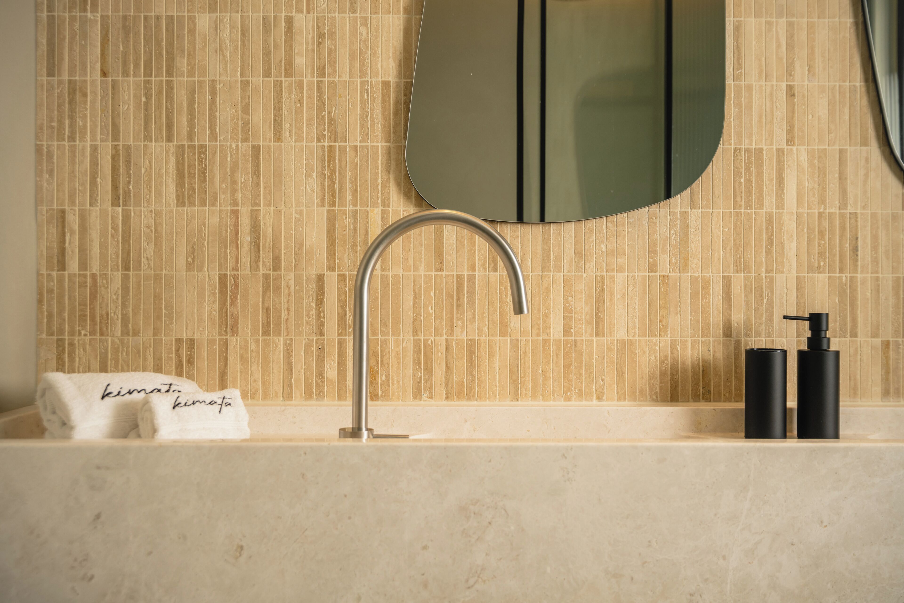 A bathroom with a tiled wall, a sink with a modern faucet, a towel with text, and two black bottles.