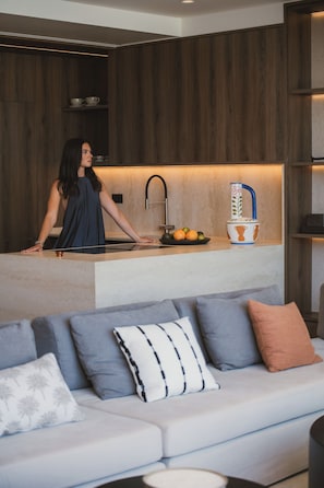 A modern kitchen with a woman standing at the counter, a bowl of fruit on the counter, and a sofa with cushions in the foreground. - Espera Beachfront Residence, Coastal City Living (Rethymno)