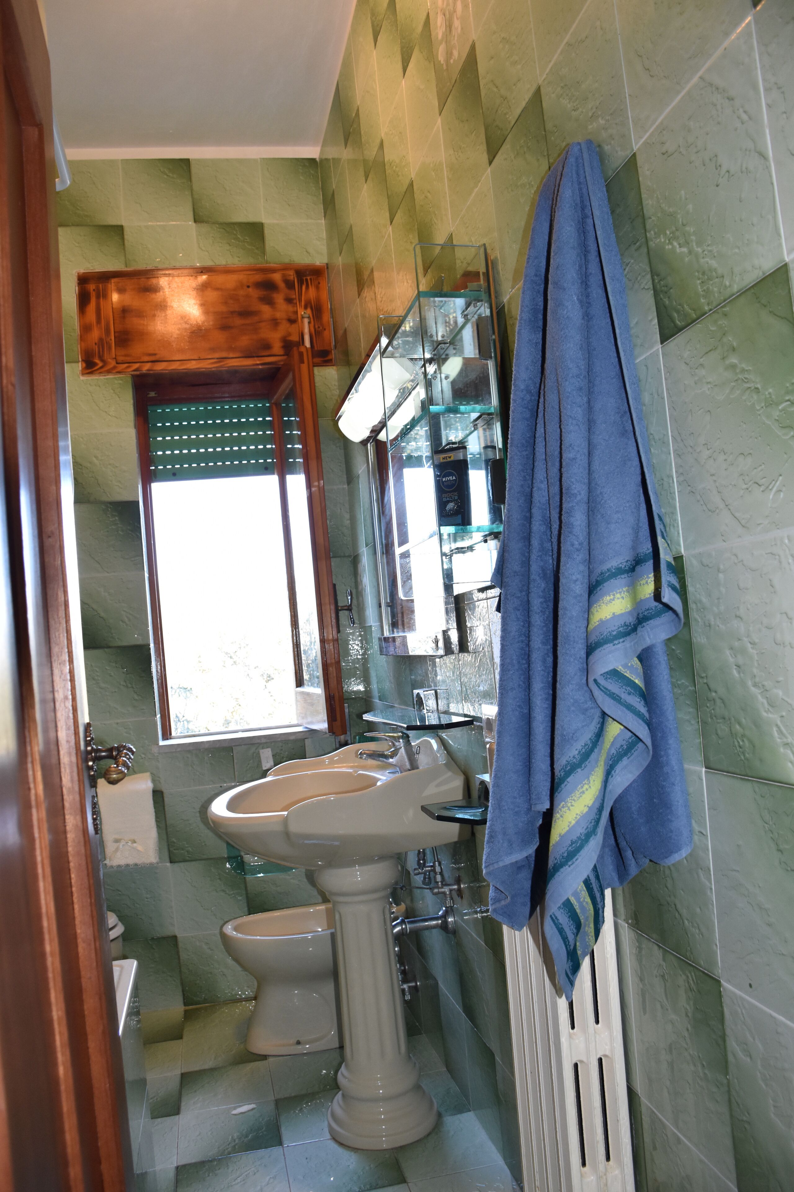 Une salle de bain avec un lavabo sur colonne, des toilettes et une fenêtre avec un store en bois.