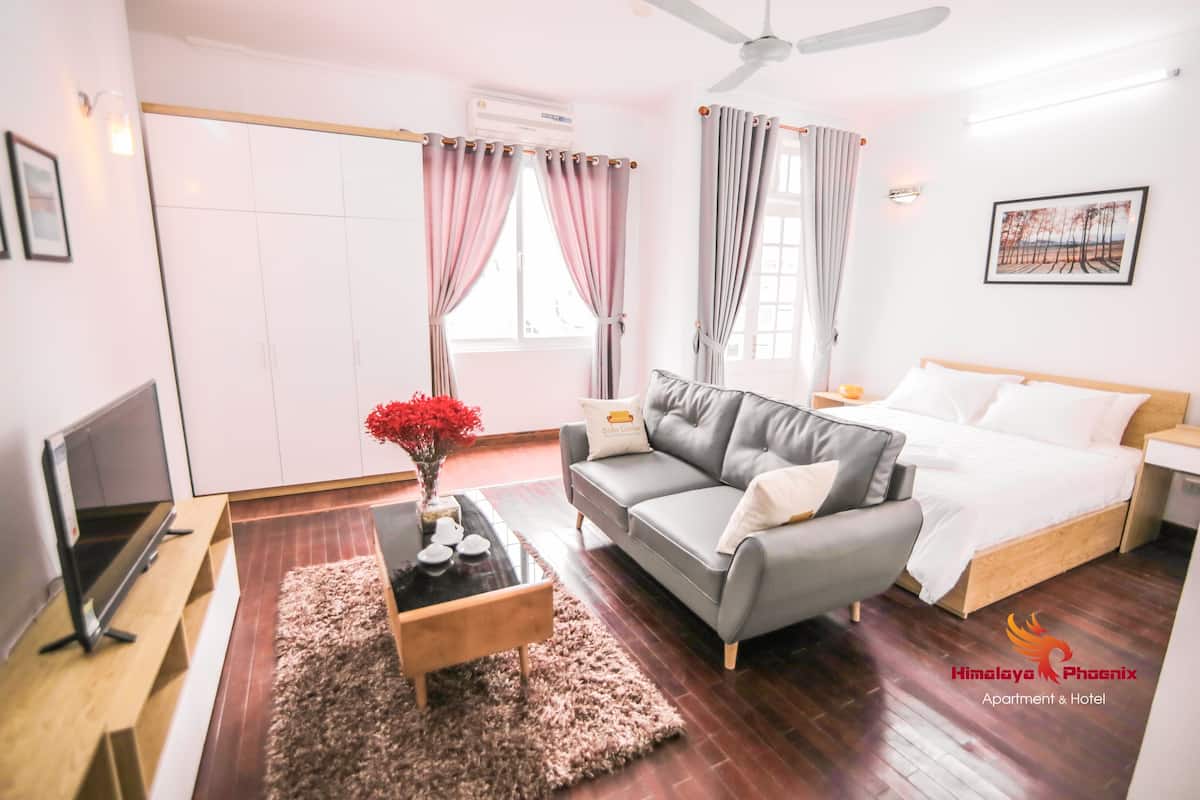 A modern hotel room with a grey sofa, a bed with white bedding, a wooden coffee table with a vase of red flowers, and a flat-screen TV on a wooden stand.