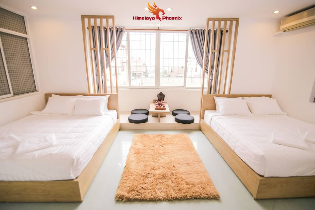 A hotel room with two beds, a wooden floor, a rug, a small table with a decorative item, and a window with curtains.