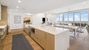 A modern kitchen with a large island, wooden cabinets, and a view of the ocean through large windows.