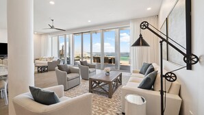 A spacious living room with a large window overlooking a beach, furnished with sofas and a coffee table.