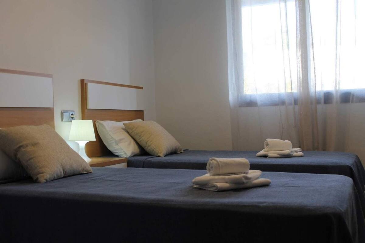 A hotel room with two beds, each with a dark blue bedspread and a rolled towel on top. There is a nightstand with a lamp and a power outlet. The room has a large window with sheer curtains.