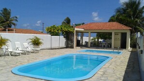 En swimmingpool med flisebelagt område, liggestole og et overd ækket siddeområde.