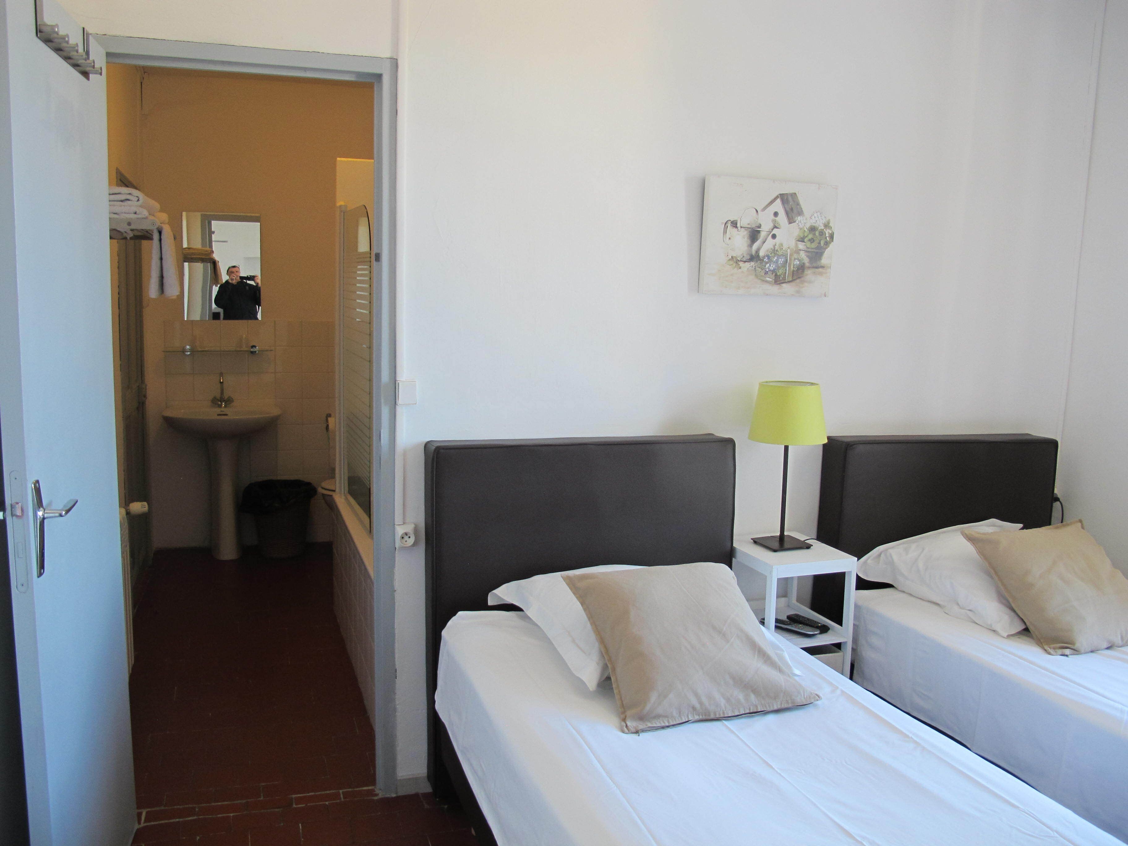 A hotel room with two beds, a bedside table with a lamp, a painting on the wall, and a bathroom visible through an open door.