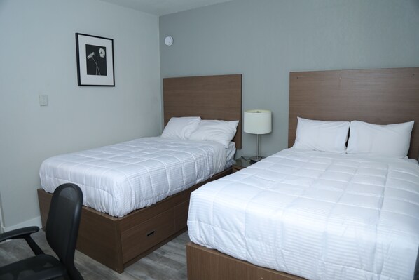 A hotel room with two single beds, a desk, a chair, a bedside lamp, and a framed picture on the wall. - Fraser Inn (Prince George)