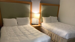A hotel room with two beds, each with white bedding and headboards. There is a bedside table with a lamp between the beds.