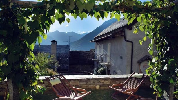 Une vue sur une montagne, avec une maison en arrière-plan. On aperçoit deux chaises en bois au premier plan.