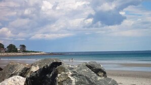 Ein Strand mit großen Felsen im Vordergrund, einem sandigen Ufer und einem ruhigen Meer unter einem teilweise bewölkten Himmel.