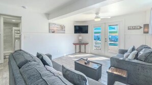 Un vaste espace de vie comprenant un canapé gris, une table basse, un téléviseur à écran plat fixé au mur et une porte donnant sur un balcon avec vue sur la piscine.