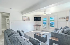 A spacious living area with a gray sofa set, a coffee table, a flat-screen TV mounted on the wall, and a door leading to a balcony with a pool view.