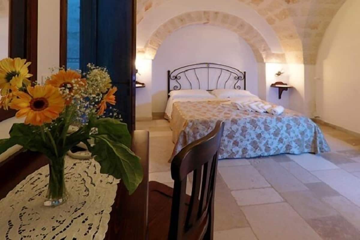 Une chambre à coucher avec un lit, une chaise, une table ornée d’un vase de fleurs et un plafond muni d’un luminaire.