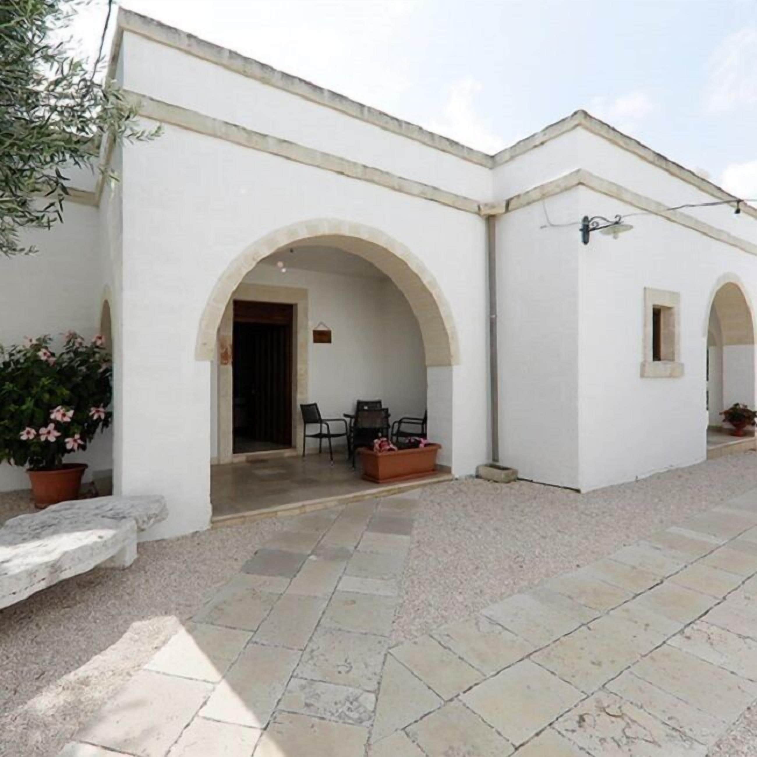 Un bâtiment blanc doté d’un patio carrelé, de deux arcades et d’un petit espace de détente avec des chaises et une table.