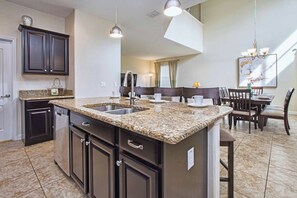 A kitchen with dark cabinetry, granite countertops, a double sink, and a dining area with a table and chairs. - 5BR Disney Pool Home | 3 King Bedrooms | Private Pool, Spa & BBQ | Sleeps 13 (Kissimmee)