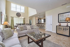 A spacious living room with a sofa, a glass coffee table, and a dining area with a chandelier. - 5BR Disney Pool Home | 3 King Bedrooms | Private Pool, Spa & BBQ | Sleeps 13 (Kissimmee)