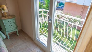 Un balcon avec une balustrade en métal, une commode verte à tiroirs et une fenêtre donnant sur un jardin et des voitures garées.