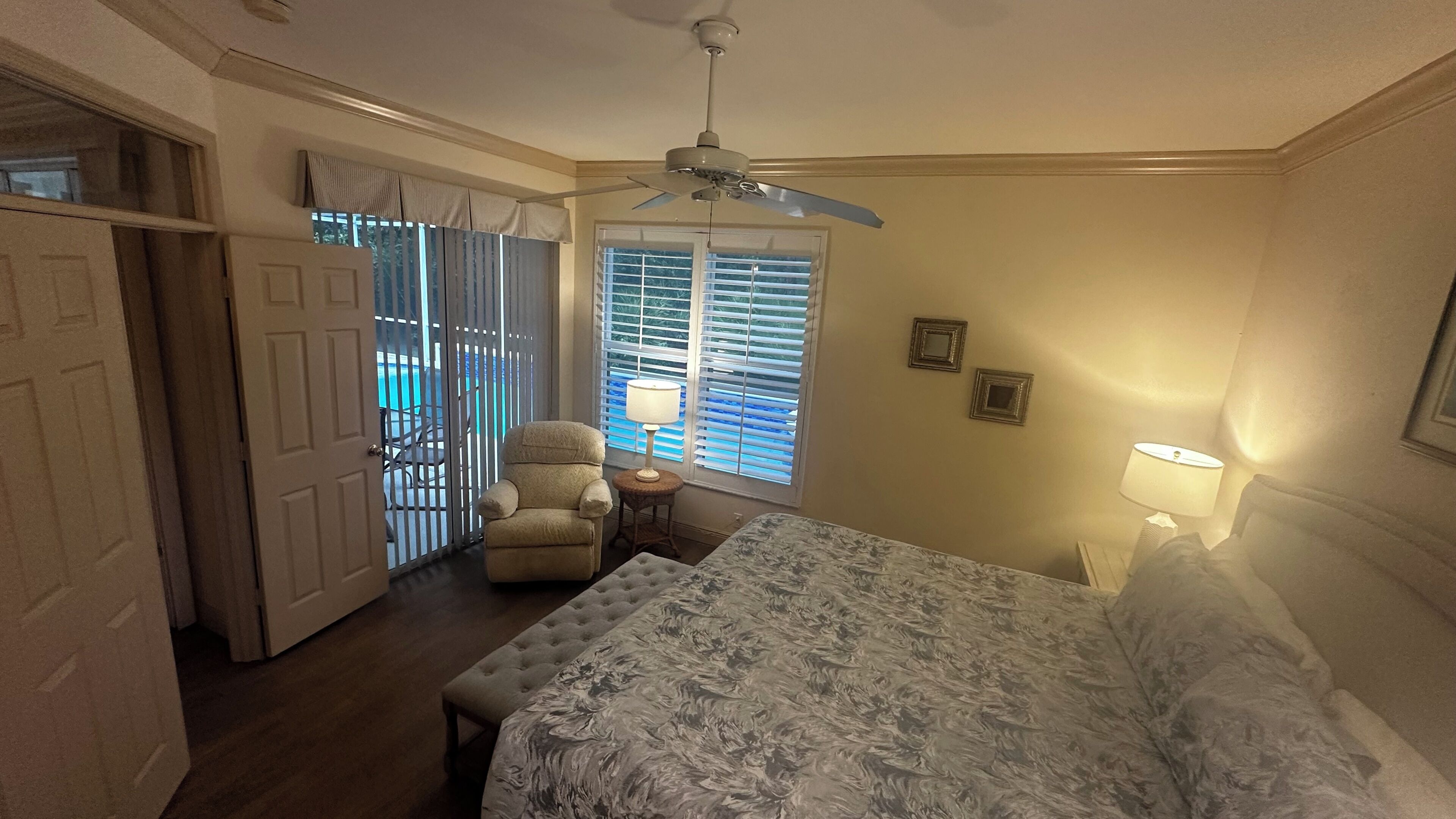 A bedroom with a bed, a chair, a window with blinds, a ceiling fan, and a door leading to a balcony.